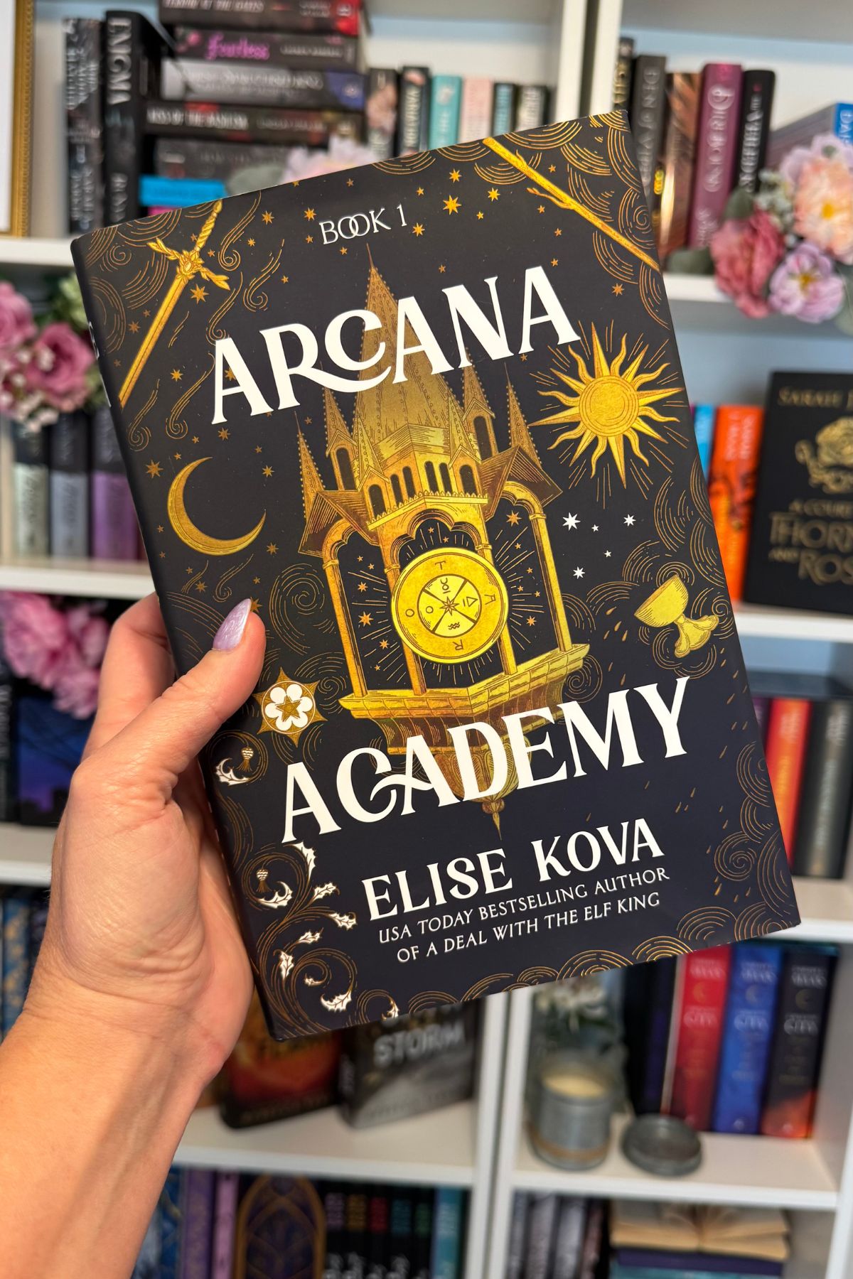 a hand holding the Arcana Academy book with bookshelf background